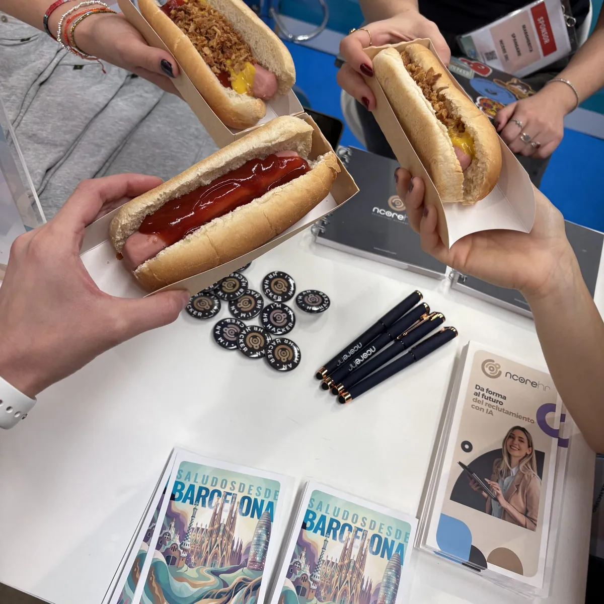 Hands of the nCore HR team holding hot dogs on the stand counter, among pins, pens, Barcelona postcards and nCore HR brochures